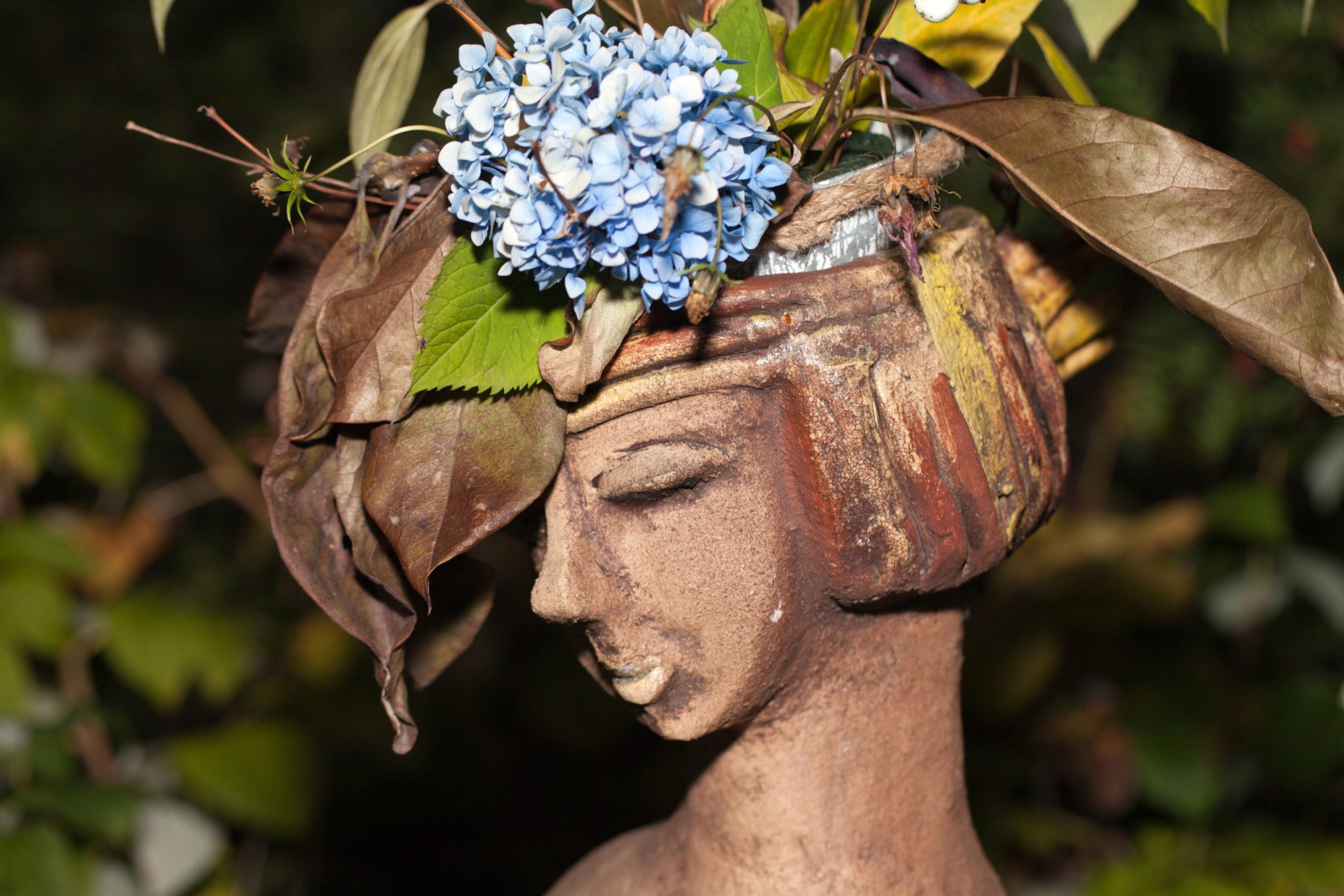 Estátua de jardim em pedra com hortênsias azuis e folhas secas pousadas na cabeça/home/klevieco/fontinha.klevie.com