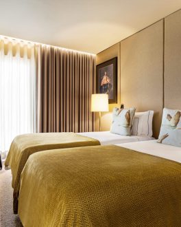 Quarto com cama de casal, colcha mostarda sobre roupa branca e almofadas, janela com cortinados claros, cadeira verde e soalho de madeira/home/klevieco/fontinha.klevie.com