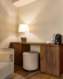 Zona de trabalho de uma suite: secretária de madeira com candeeiro e máquina de café, poltrona clara e pufe redondo, paredes em tom creme/home/klevieco/fontinha.klevie.com