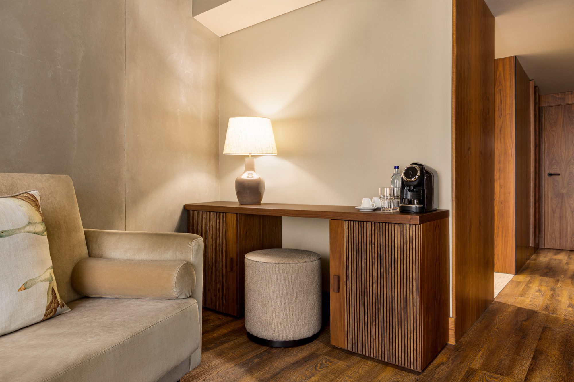 Zona de trabalho de uma suite: secretária de madeira com candeeiro e máquina de café, poltrona clara e pufe redondo, paredes em tom creme/home/klevieco/fontinha.klevie.com