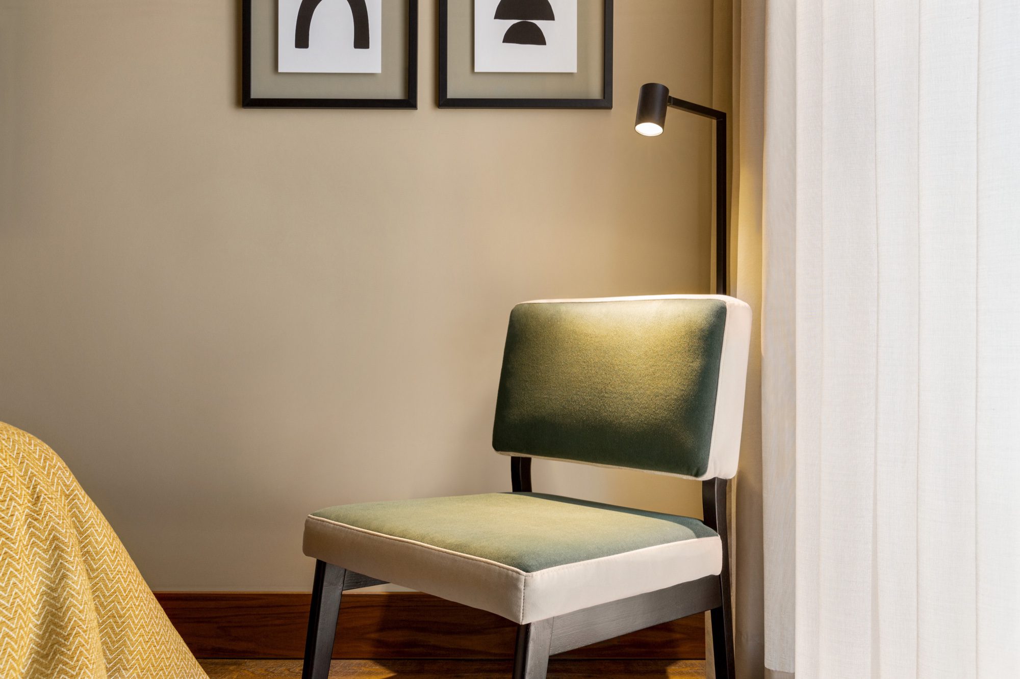 Cadeira estofada a verde junto à janela de um quarto, com dois quadros a preto e branco na parede e a ponta da cama coberta por uma manta mostarda/home/klevieco/fontinha.klevie.com