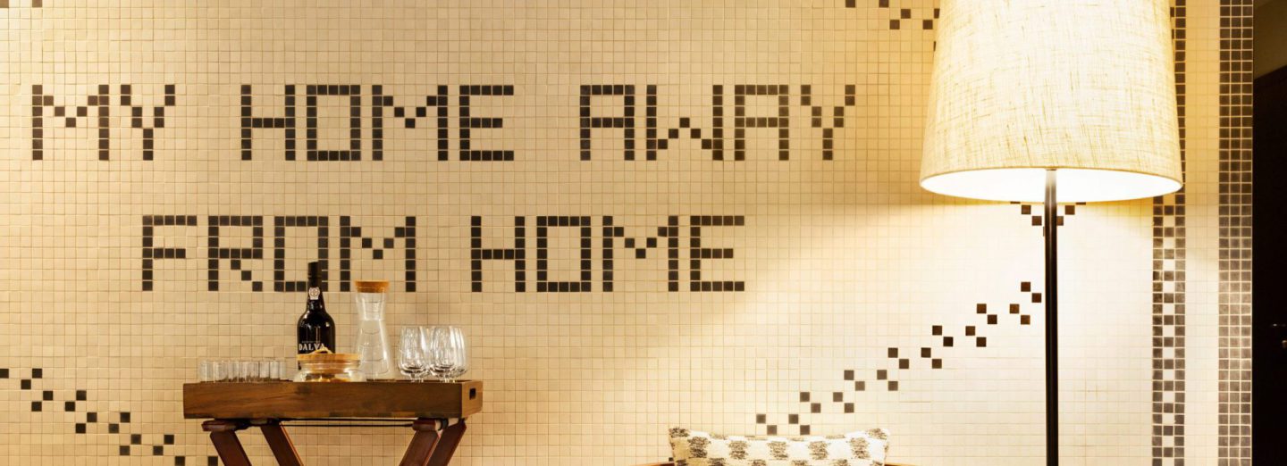 Parede de azulejos dourados com a frase «MY HOME AWAY FROM HOME» desenhada a mosaico escuro, com um candeeiro de pé e uma mesa de apoio com copos/home/klevieco/fontinha.klevie.com