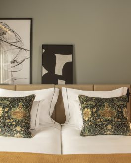 Cabeceira de cama com duas almofadas de padrão floral escuro sobre roupa branca, entre candeeiros de mesa e quadros numa parede verde-acinzentada/home/klevieco/fontinha.klevie.com